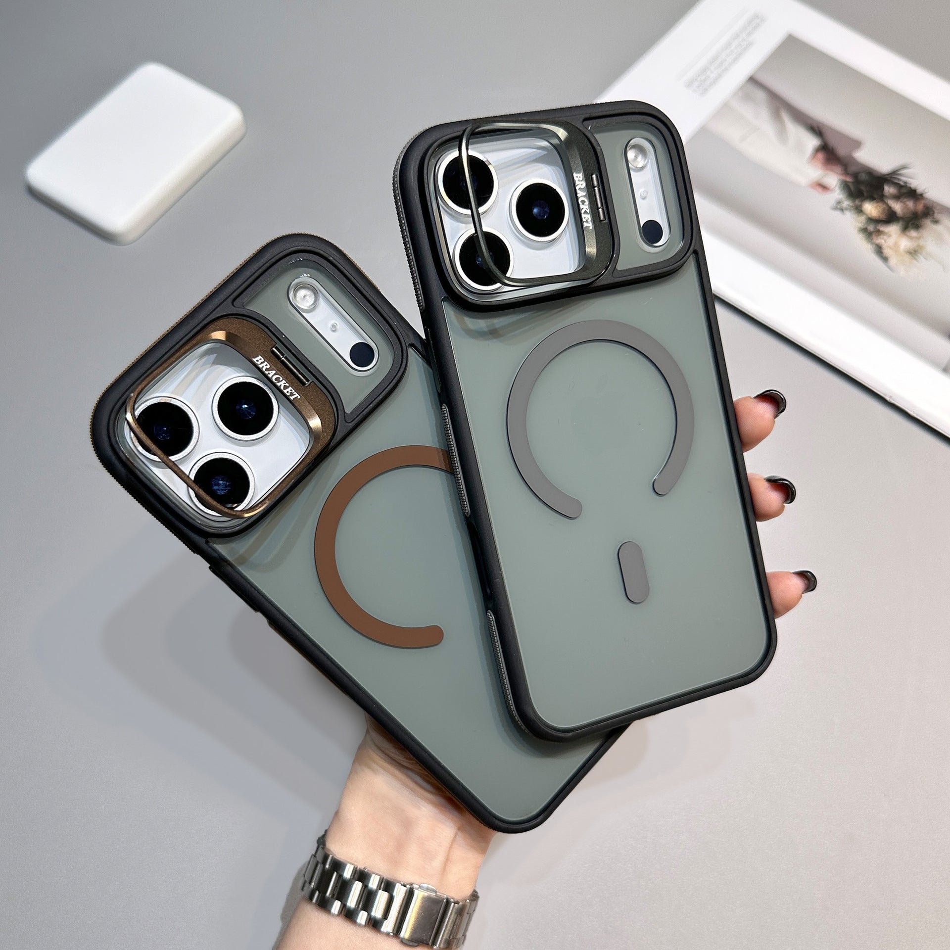 CaseSun-Hand holding two iPhone 17 Pro Max cases with matte translucent backs and built-in magnetic rings, shown in bronze and black lens ring colors.